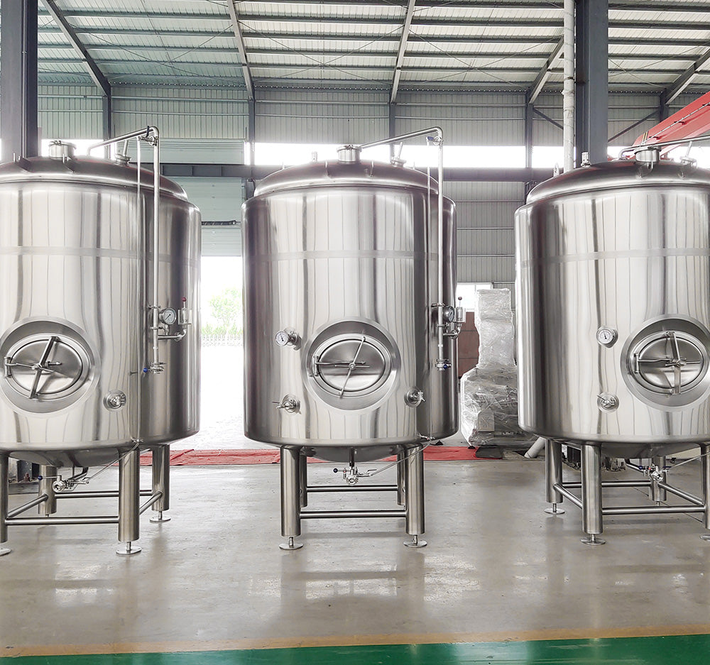 Three large stainless steel beer tanks standing on a concrete floor inside a warehouse or brewery facility.