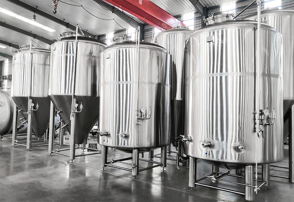 Large stainless steel tanks in a brewery interior with concrete floors and industrial ceiling structures.