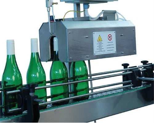 A stainless steel industrial bottle-capping machine processing four green glass bottles on a conveyor belt, shown against a white background.