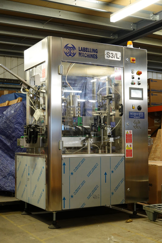 A stainless steel continuous bottle labeller machine with a glass front door, photographed in a warehouse setting.